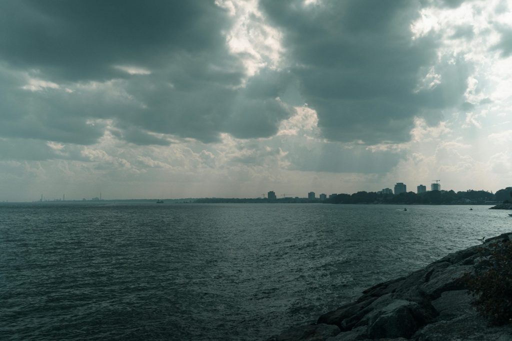 A serene lakeshore landscape with dramatic cloud formations and city silhouette in the background.