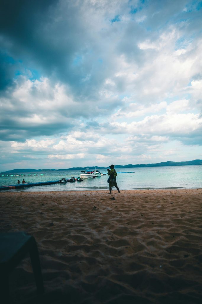 A vibrant beach setting in Phuket, showing a lone walker on the sand under a dramatic cloudy sky.